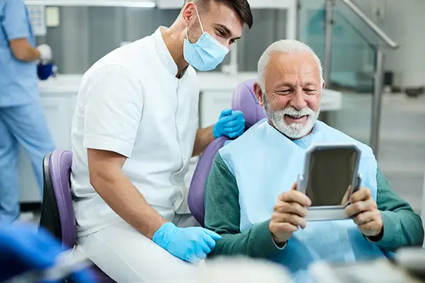 Happy, senior patient looking at himself with all on x dental implants in a mirror at Surprise Oral & Implant Surgery in Surprise, AZ